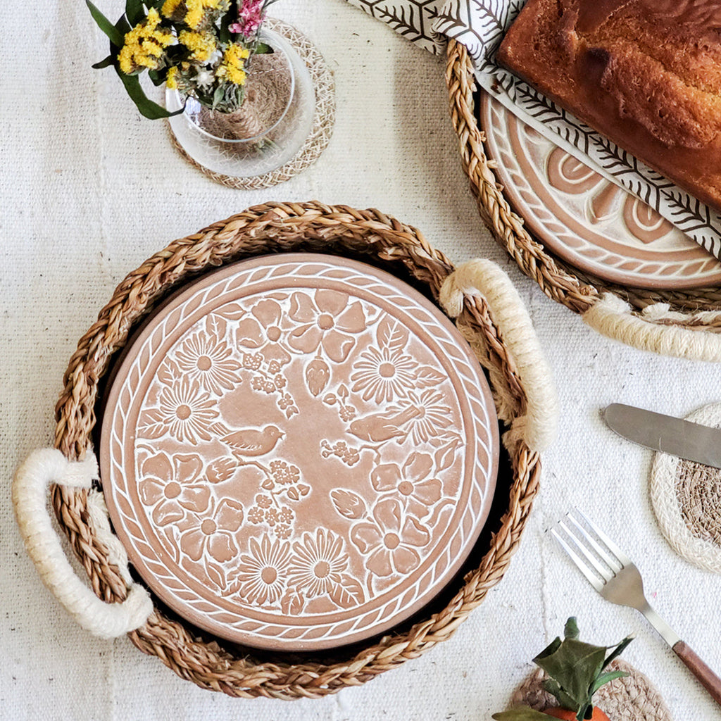 Terra cotta warmer plate with floral and bird pattern in a hand woven bread basket surrounded by tableware on a linen tablecloth.