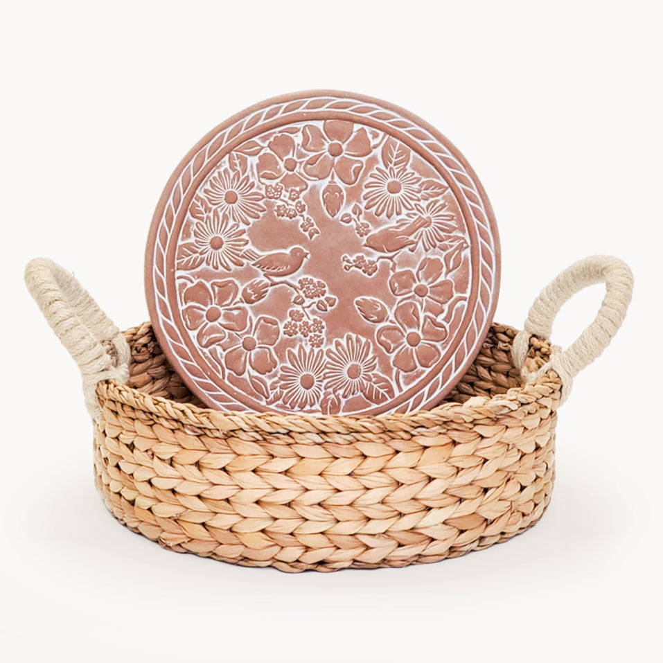 Terra cotta warmer plate with floral bird patterns in a hand woven bread basket on a white background