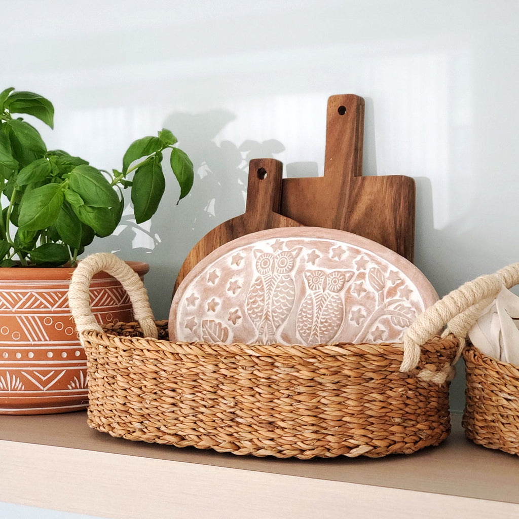 Woven bread basket with terra cotta warmer plate and wooden cutting boards on a kitchen shelf with basil in the background.