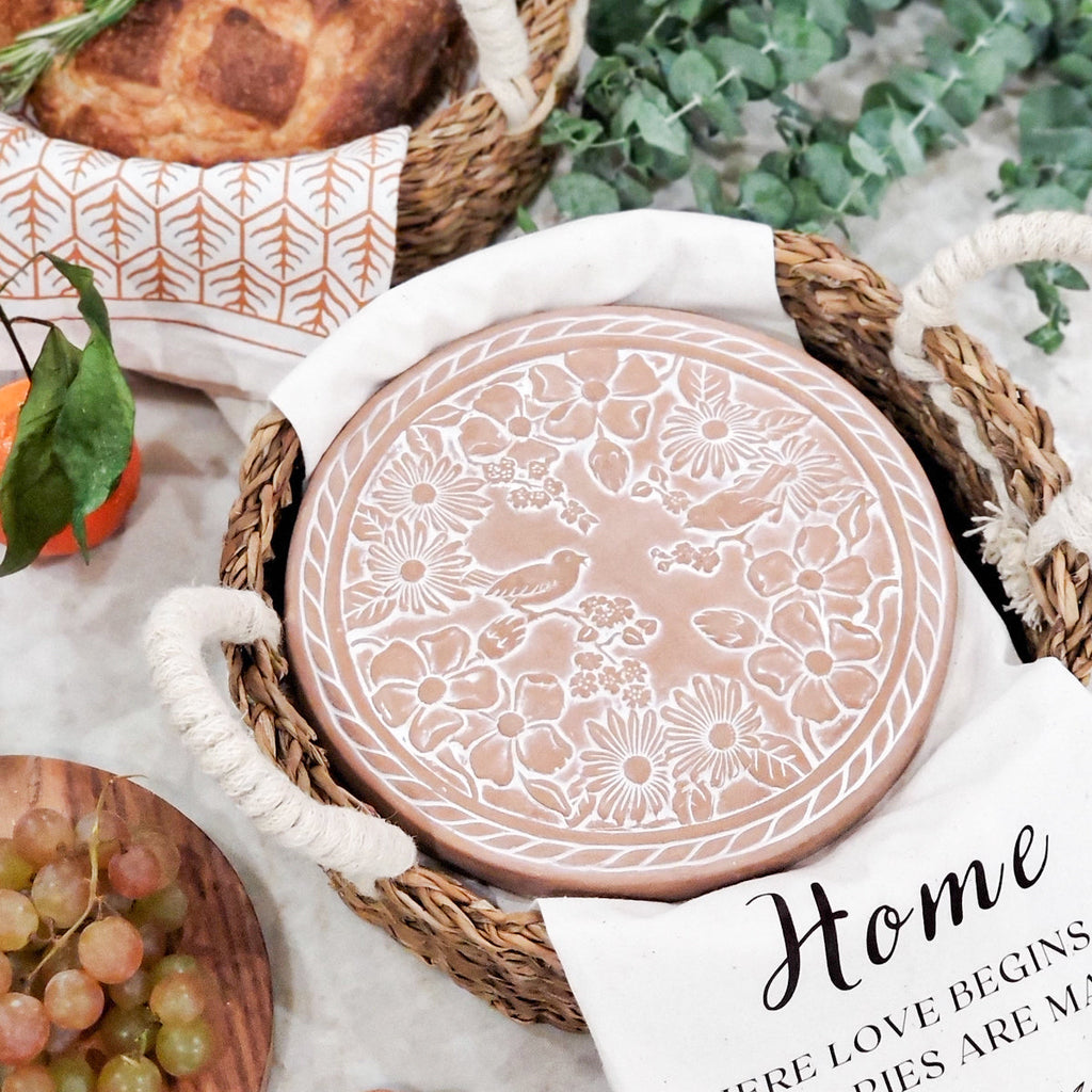 Terra cotta warmer plate with floral bird pattern in a woven bread basket with crusty bread, grapes, and a kitchen tea towel.