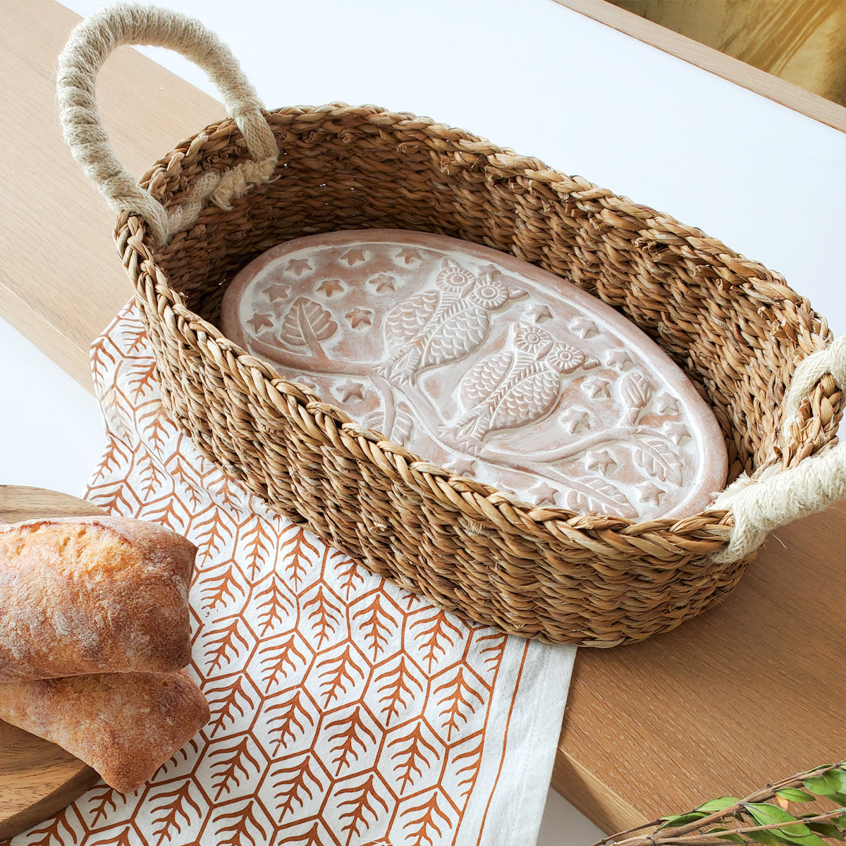 Woven bread warmer basket with terra cotta plate on a kitchen counter.