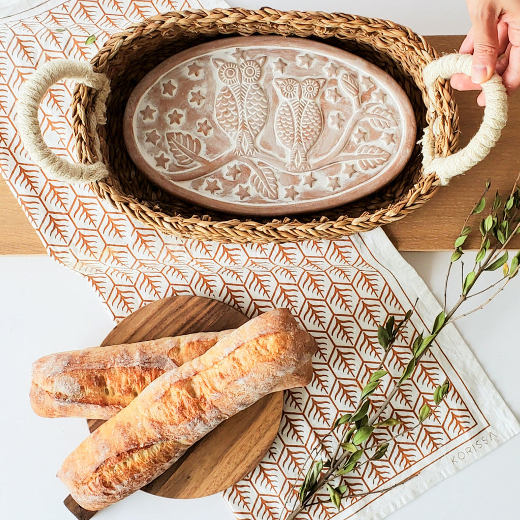 Decorative terra cotta plate with owl design in a woven bread warmer basket on kitchen counter with bread and greenery.