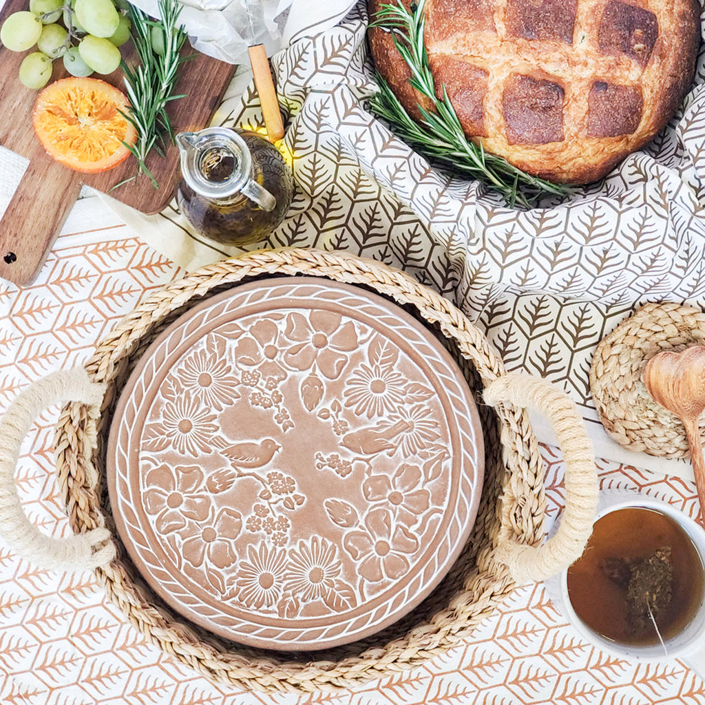 Terra cotta warmer plate with birds in a garden design in a hand woven bread basket on a patterned tablecloth with bread and fruit.