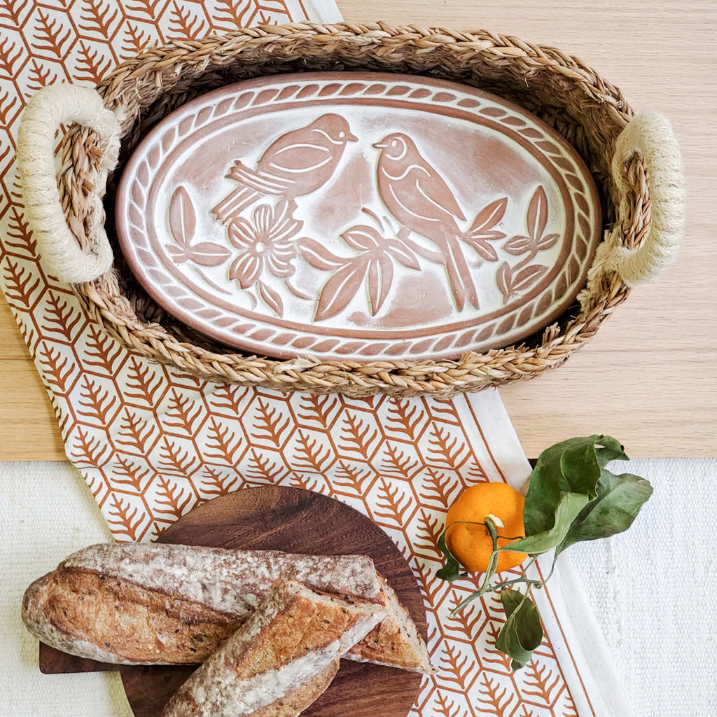 Decorative plate with bird design in a woven basket on a table with bread and an orange.