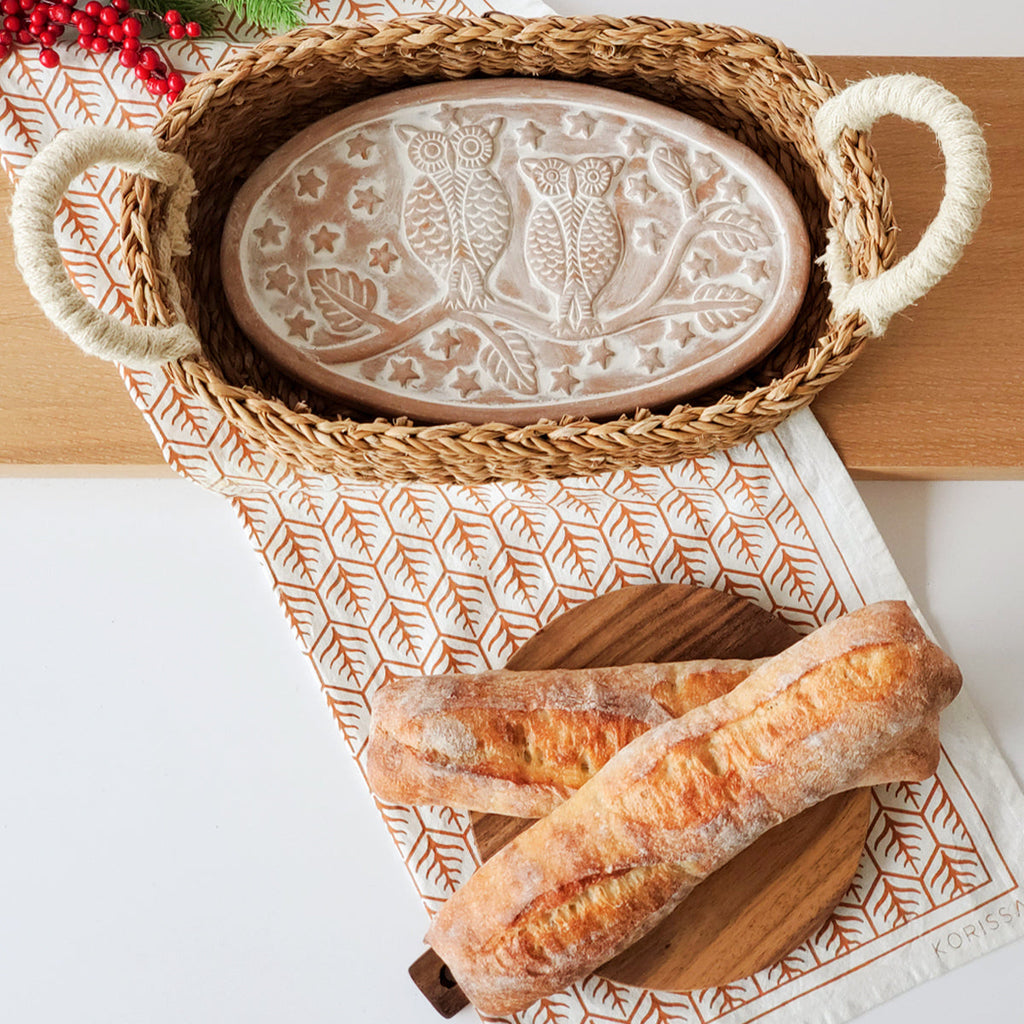 Terra Cotta warmer plate with owl design in a woven bread basket on a kitchen counter with crusty bread.