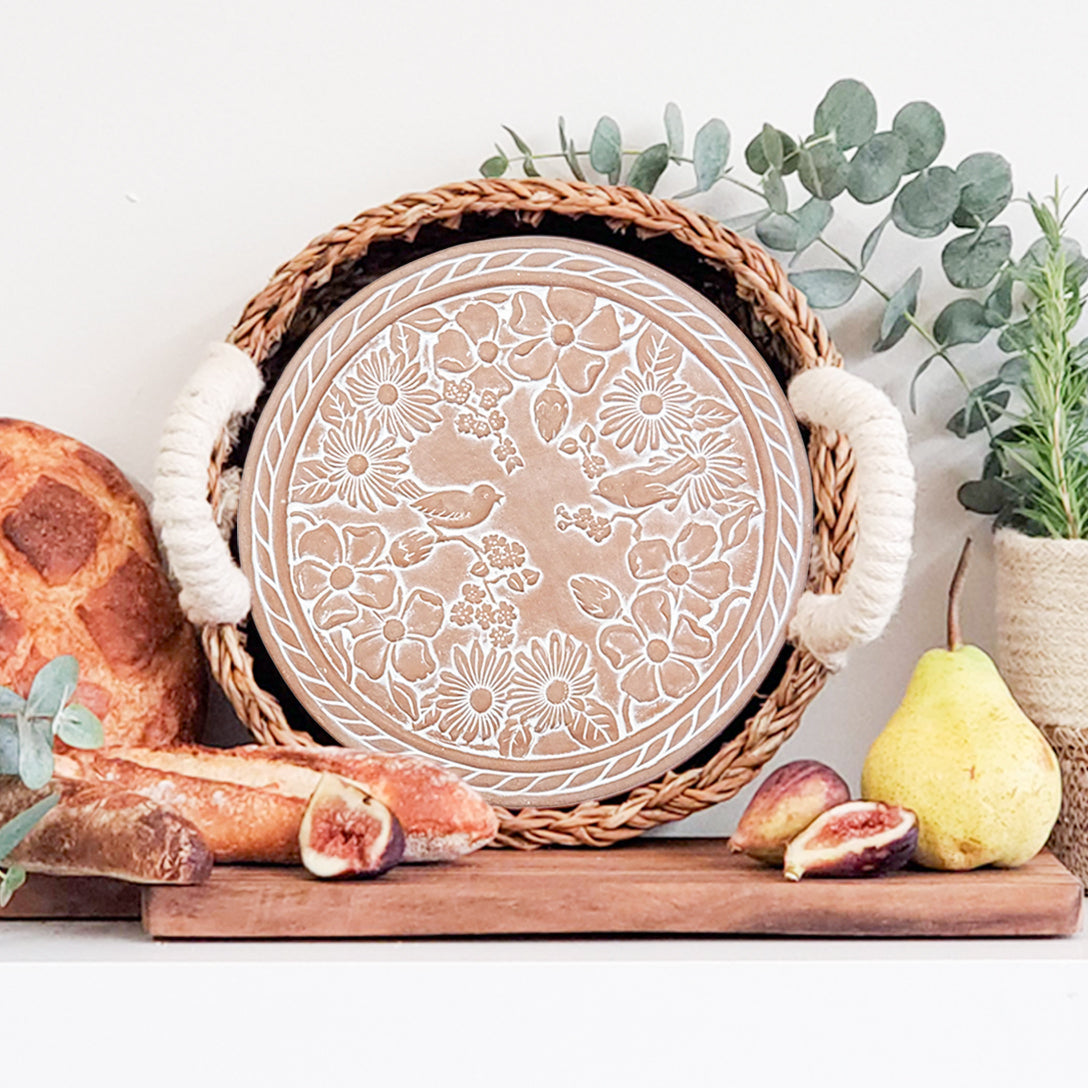 Terra cotta warmer plate with garden flower design in a hand woven bread basket, surrounded by bread, figs, and pears on a white surface.